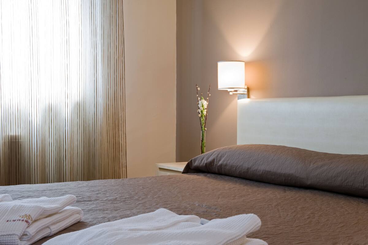 Una cama bien hecha con un cabecero marrón, una lámpara de noche y toallas dobladas sobre una mesita de noche de madera.