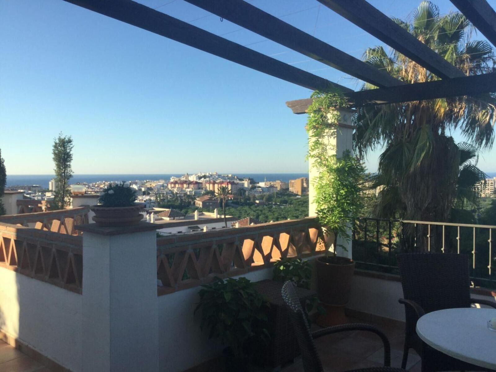 A balcony with a view of a coastal city, a table, chairs, and potted plants.
