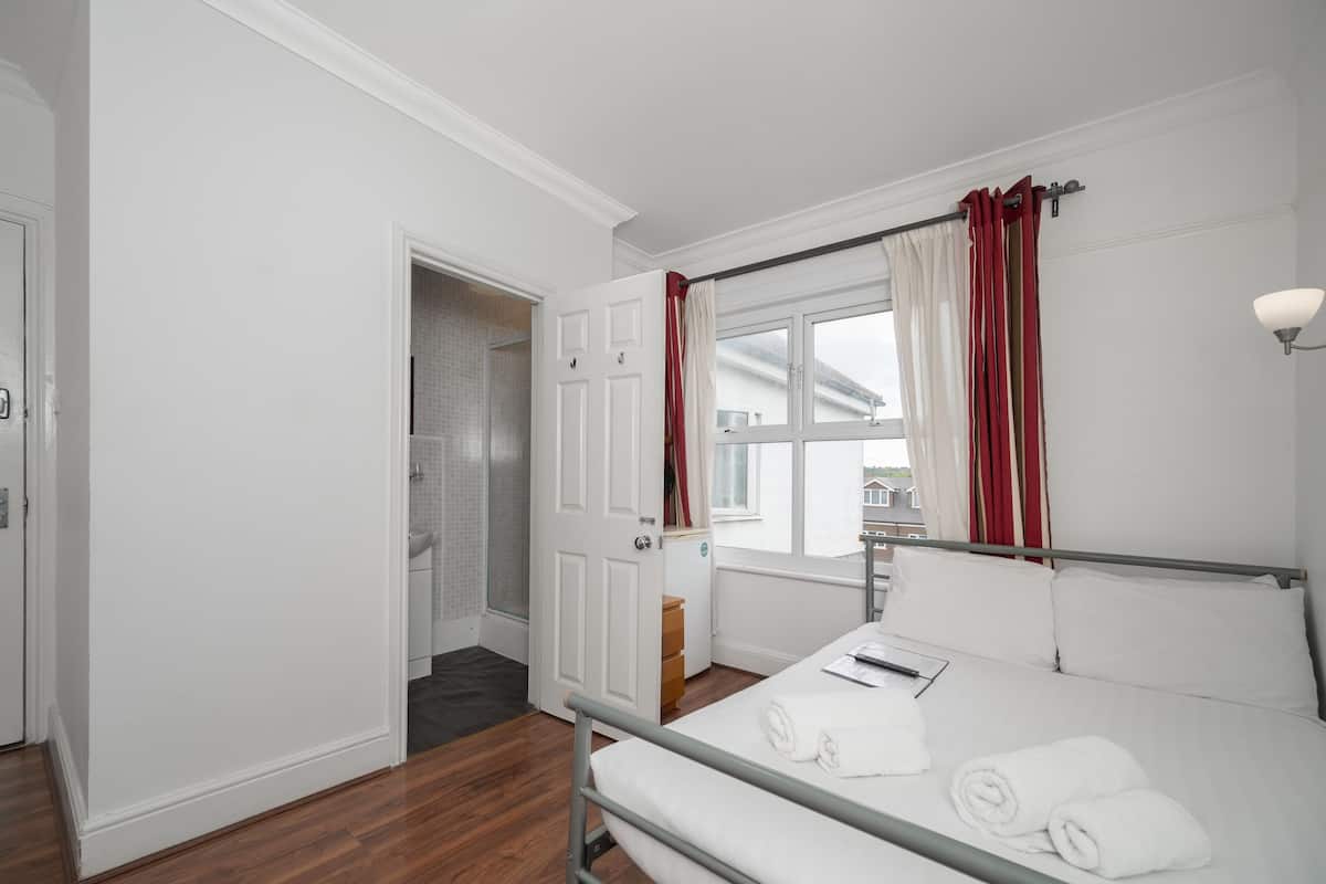 Quarto de hotel com cama, janela com cortinas vermelhas, mesa de cabeceira com abajur e vista para o exterior.