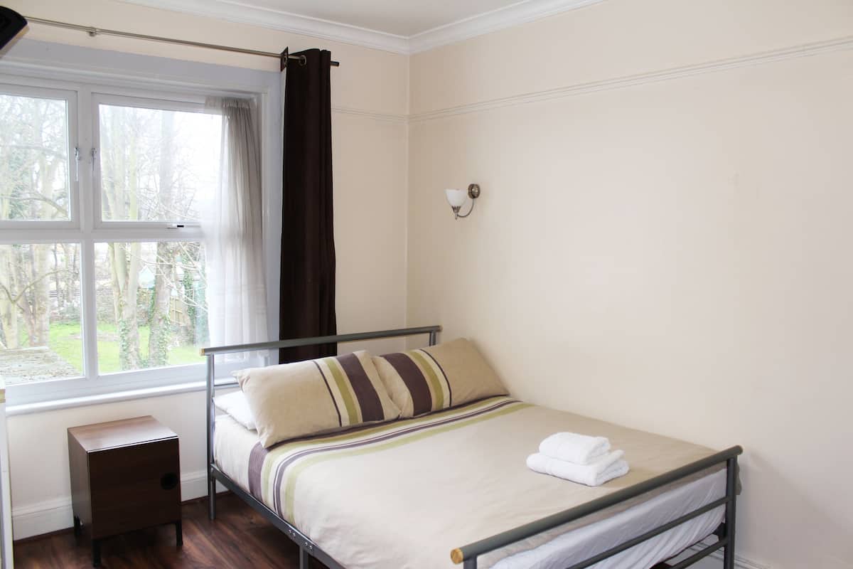 Uma cama de solteiro com travesseiro listrado, mesa de cabeceira, janela com cortinas e luminária fixada na parede.