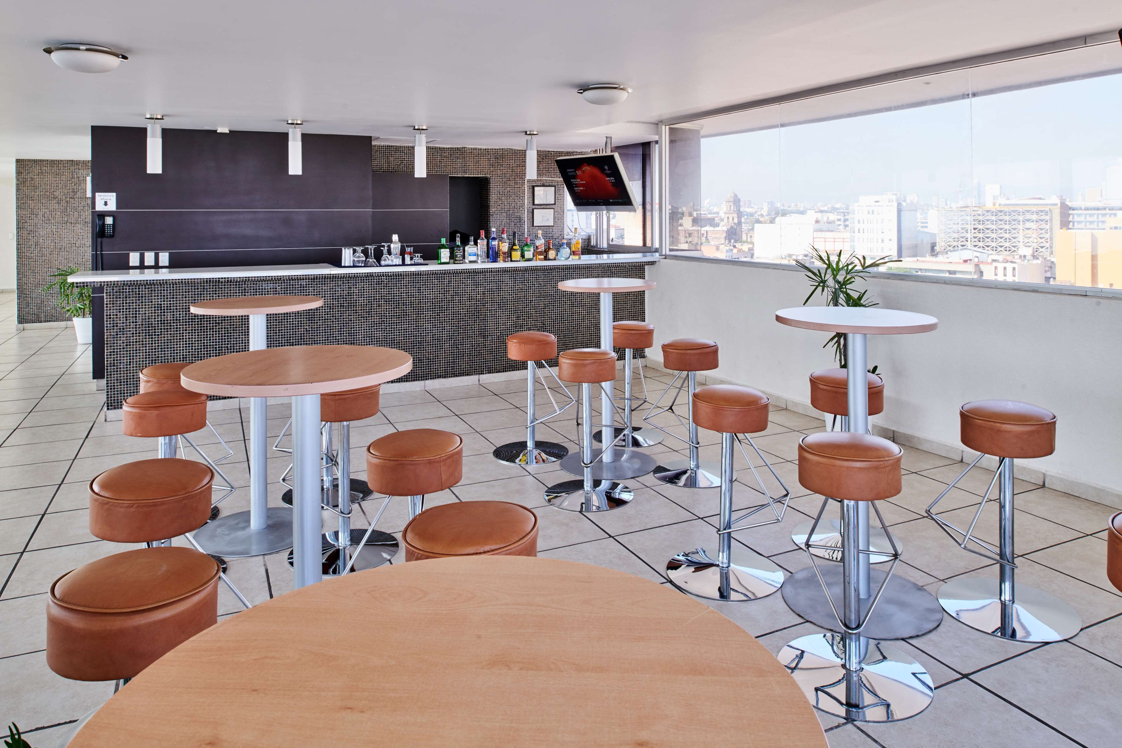 Un espace bar moderne avec vue sur la ville, comprenant des tables rondes et des tabourets, un grand écran et un sol carrelé.