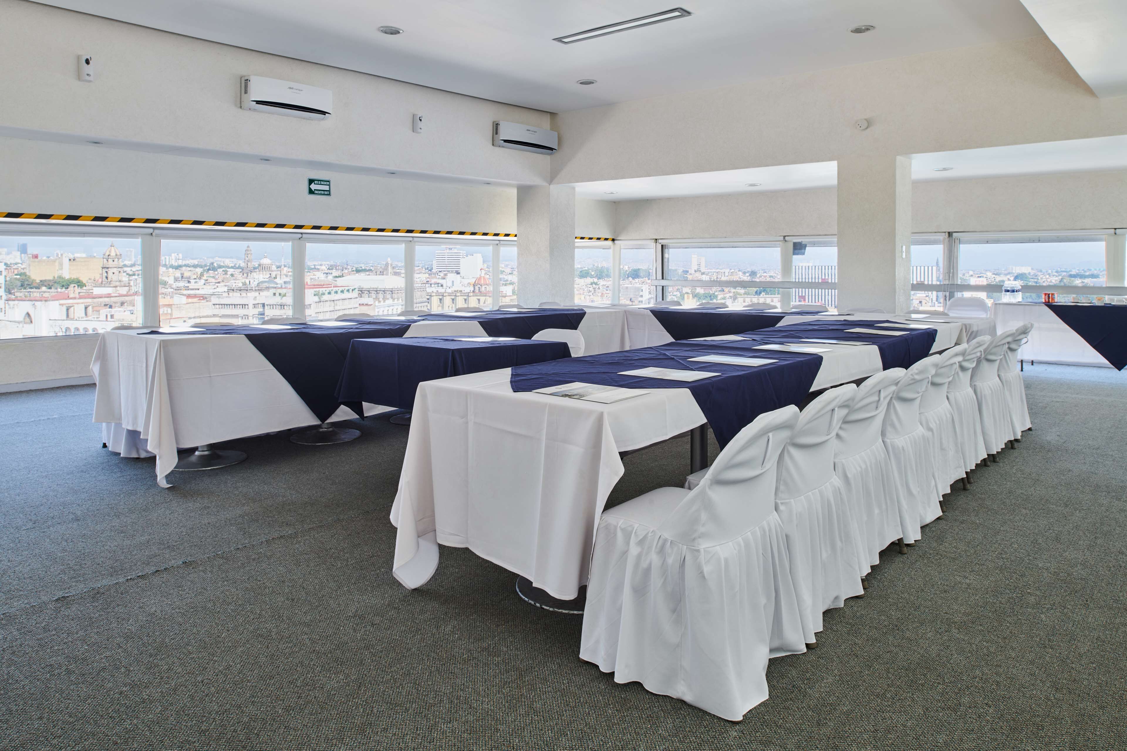A conference room with tables and chairs, large windows with a city view, and air conditioning units on the ceiling.