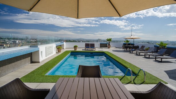 Un área de piscina en la azotea con vistas a la ciudad, sillones de descanso y sombrillas.