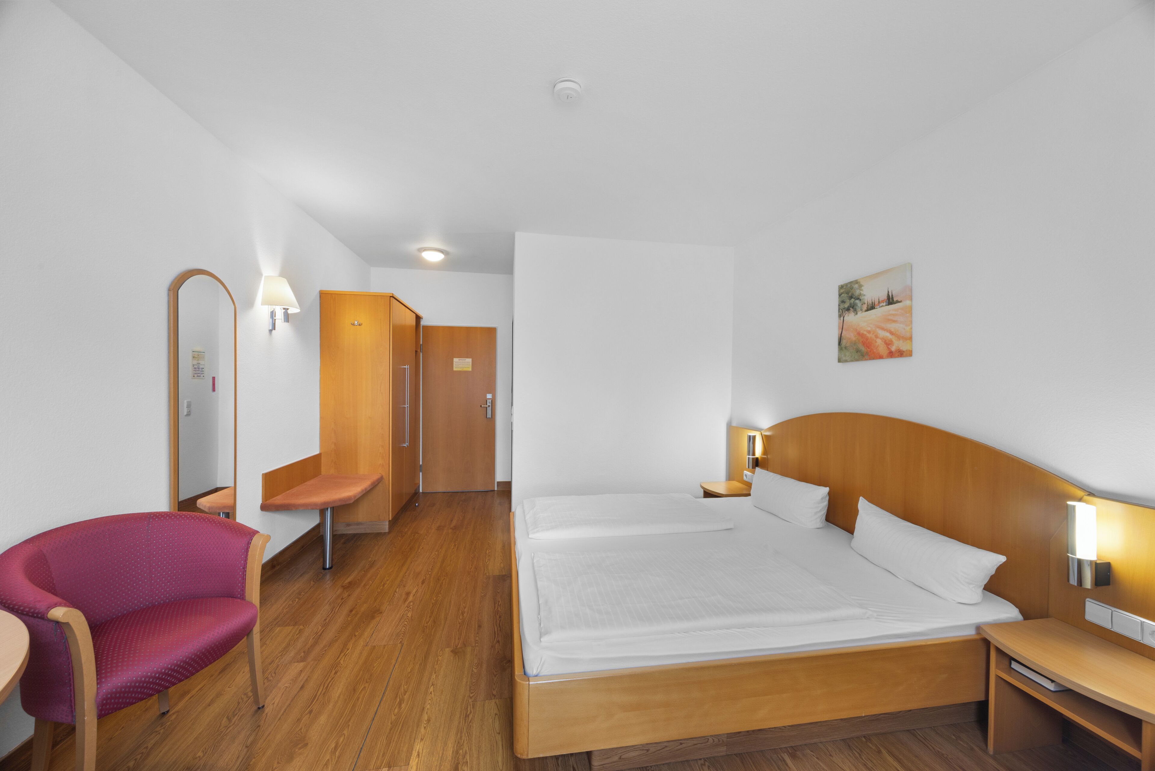 Ein Hotelzimmer mit einem großen Bett, einem Sessel, einem Schreibtisch, einem Spiegel und einer Tür.