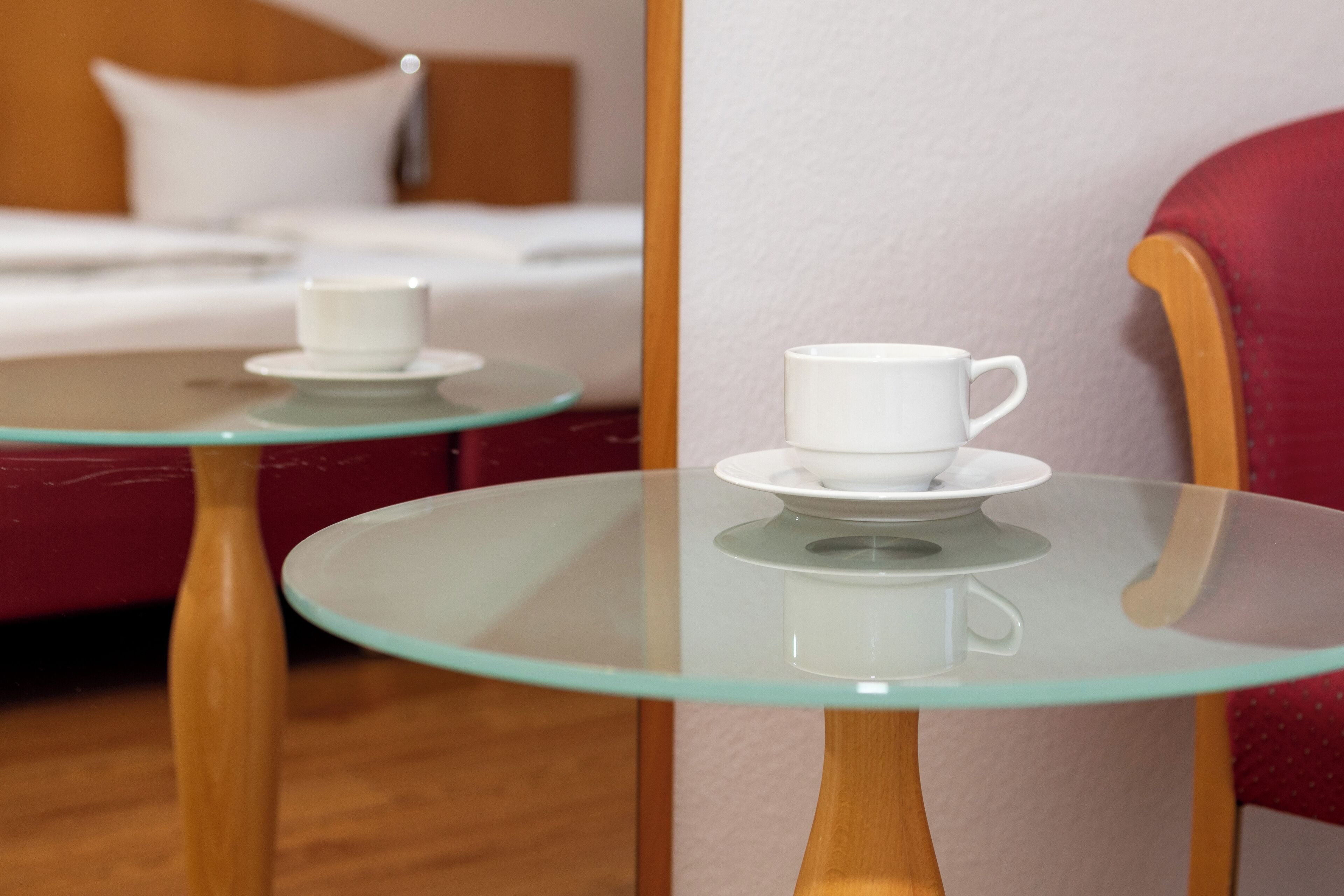 A hotel room with a bed, a red armchair, and two glass-top tables with white cups.