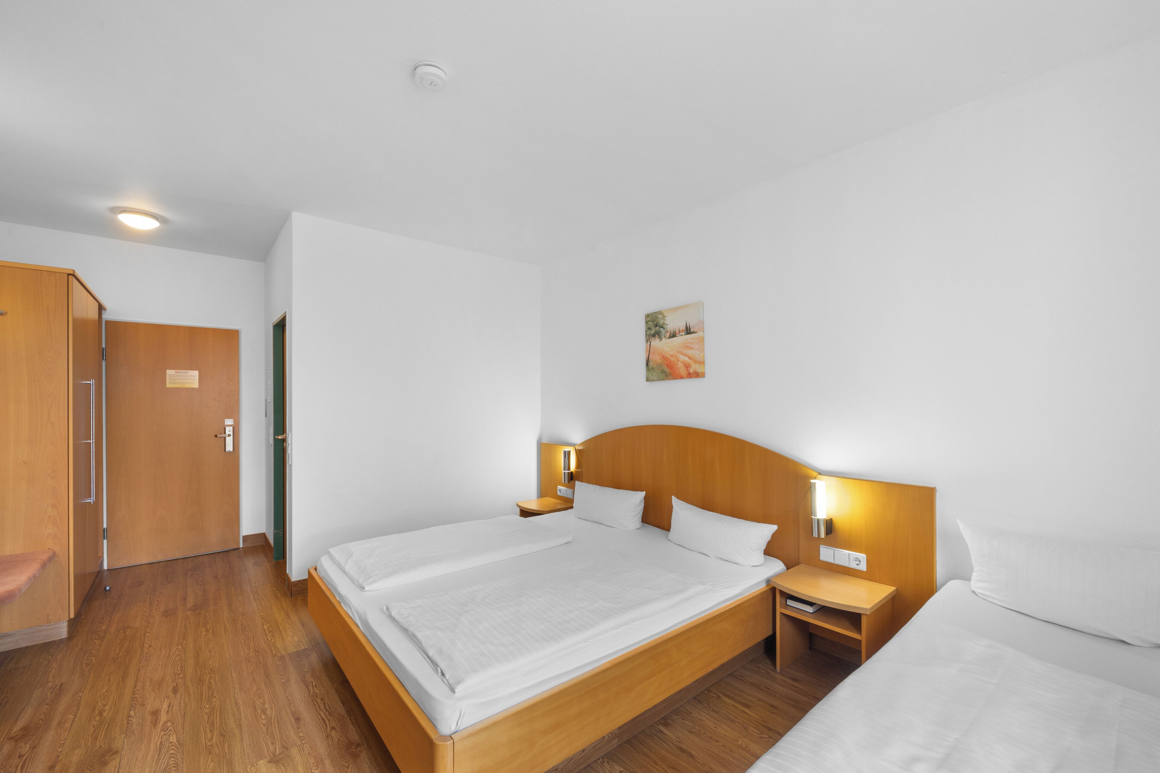 A hotel room with a wooden bed, a nightstand, a bedside table, and a door leading to another room.