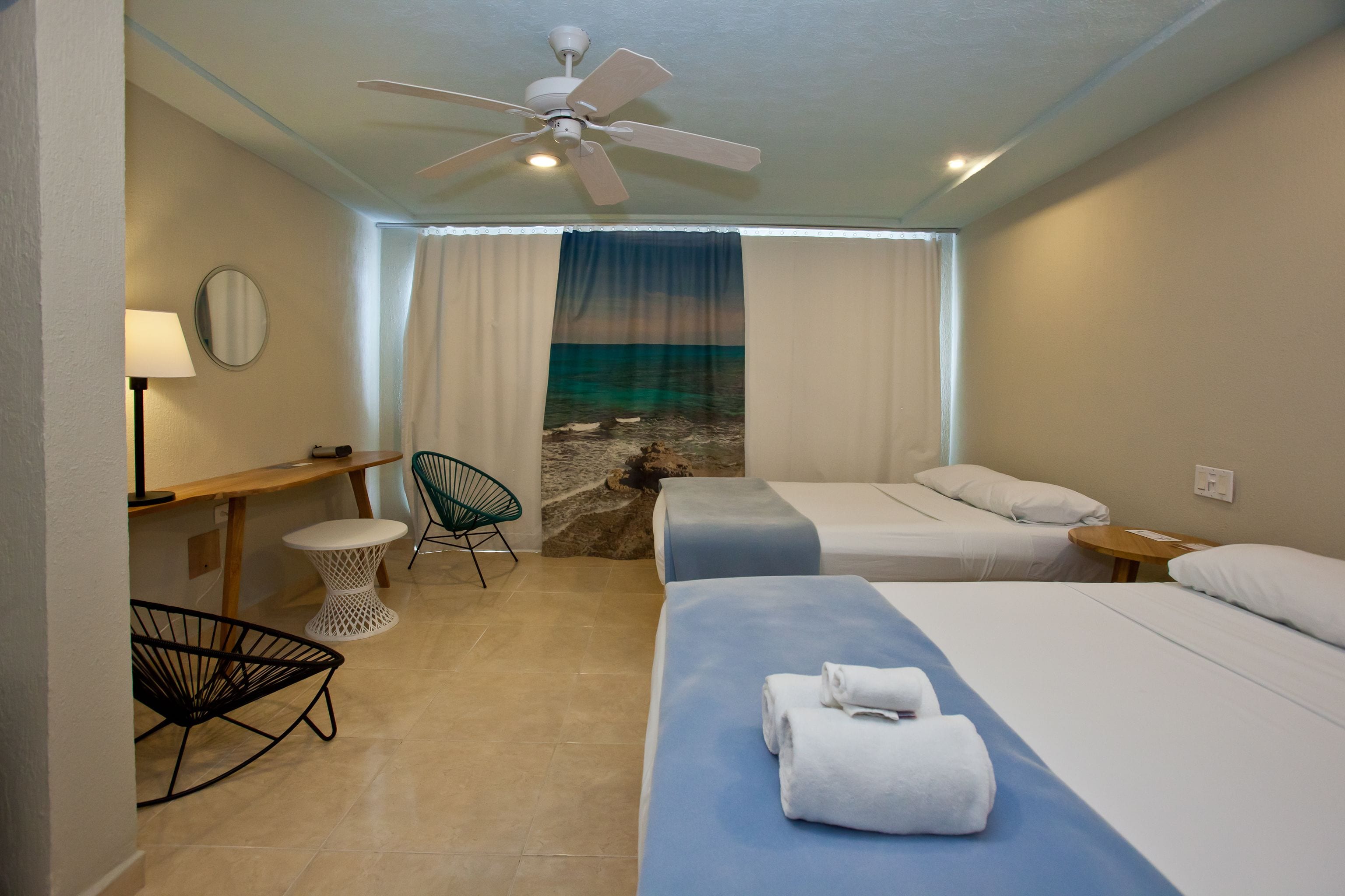 Habitación de hotel con dos camas, un ventilador de techo, una pequeña mesa con lámpara, una silla y vista a la playa a través de una ventana.