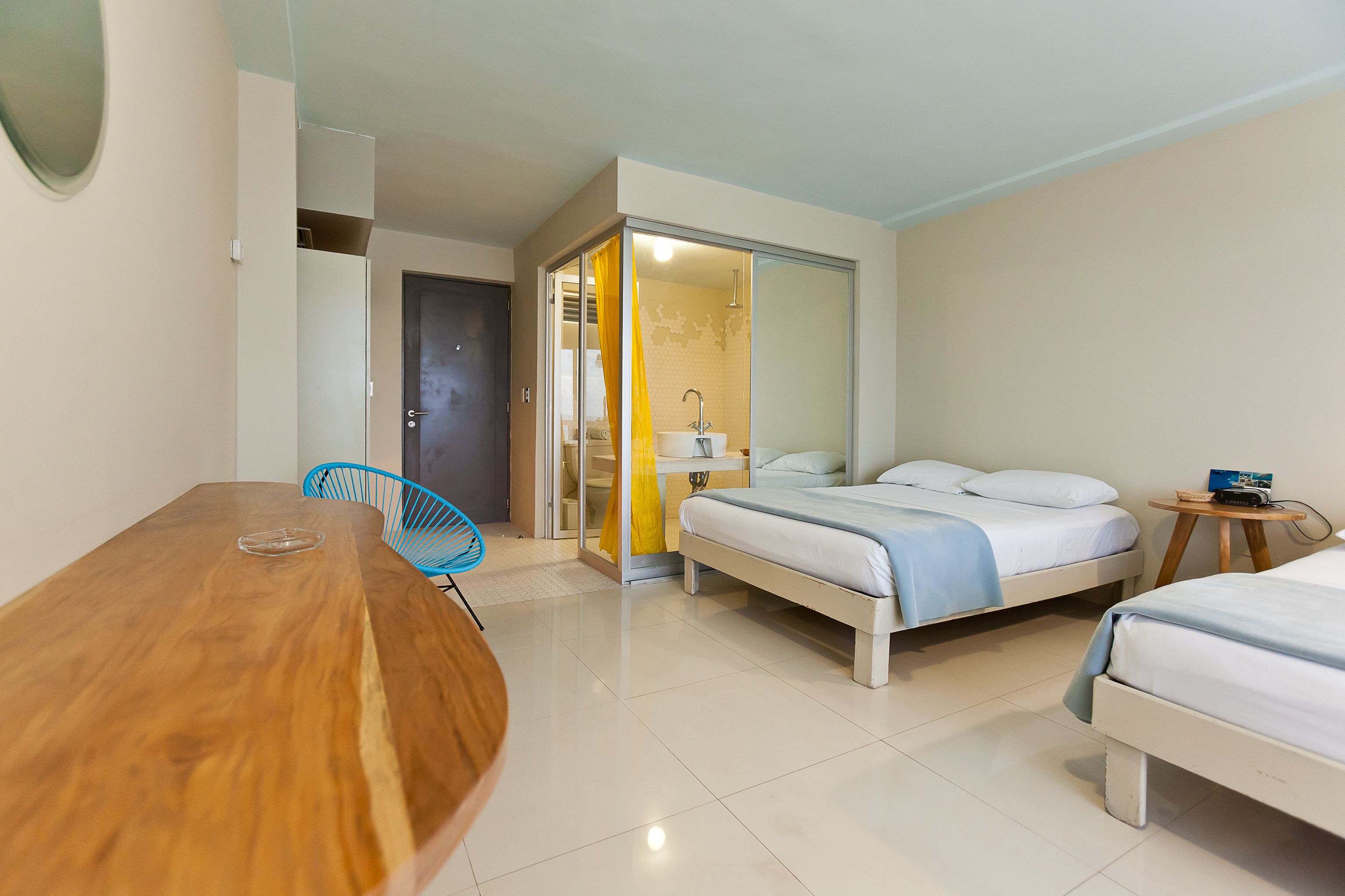 A hotel room with a wooden dining table, two single beds, a small bedside table, and a bathroom with a shower.
