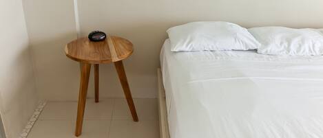 A bedroom with a bed, white pillows, a small wooden side table, and a white wall.