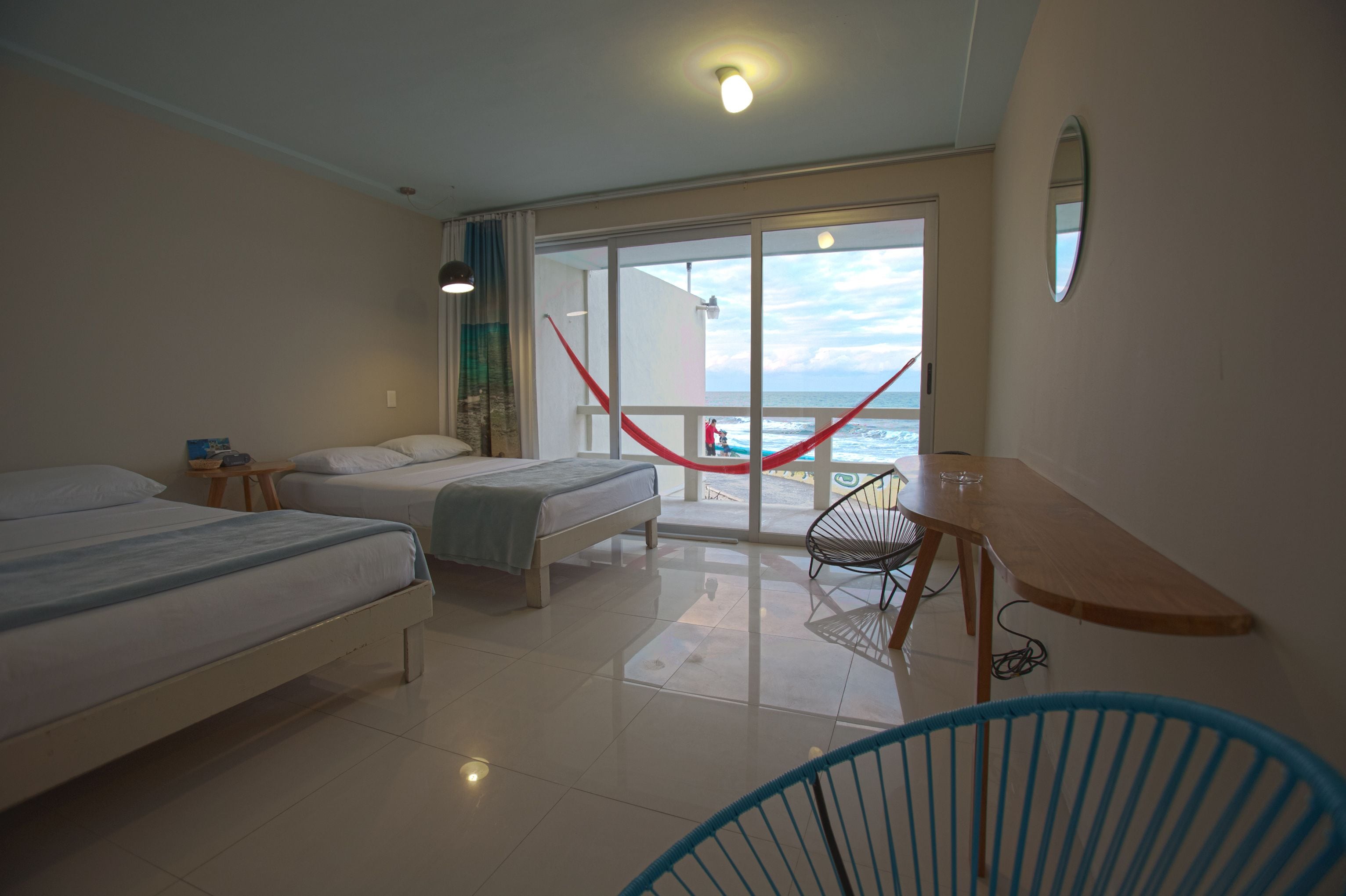 A hotel room with two beds, a small table, a chair, and a view of the ocean through large windows.
