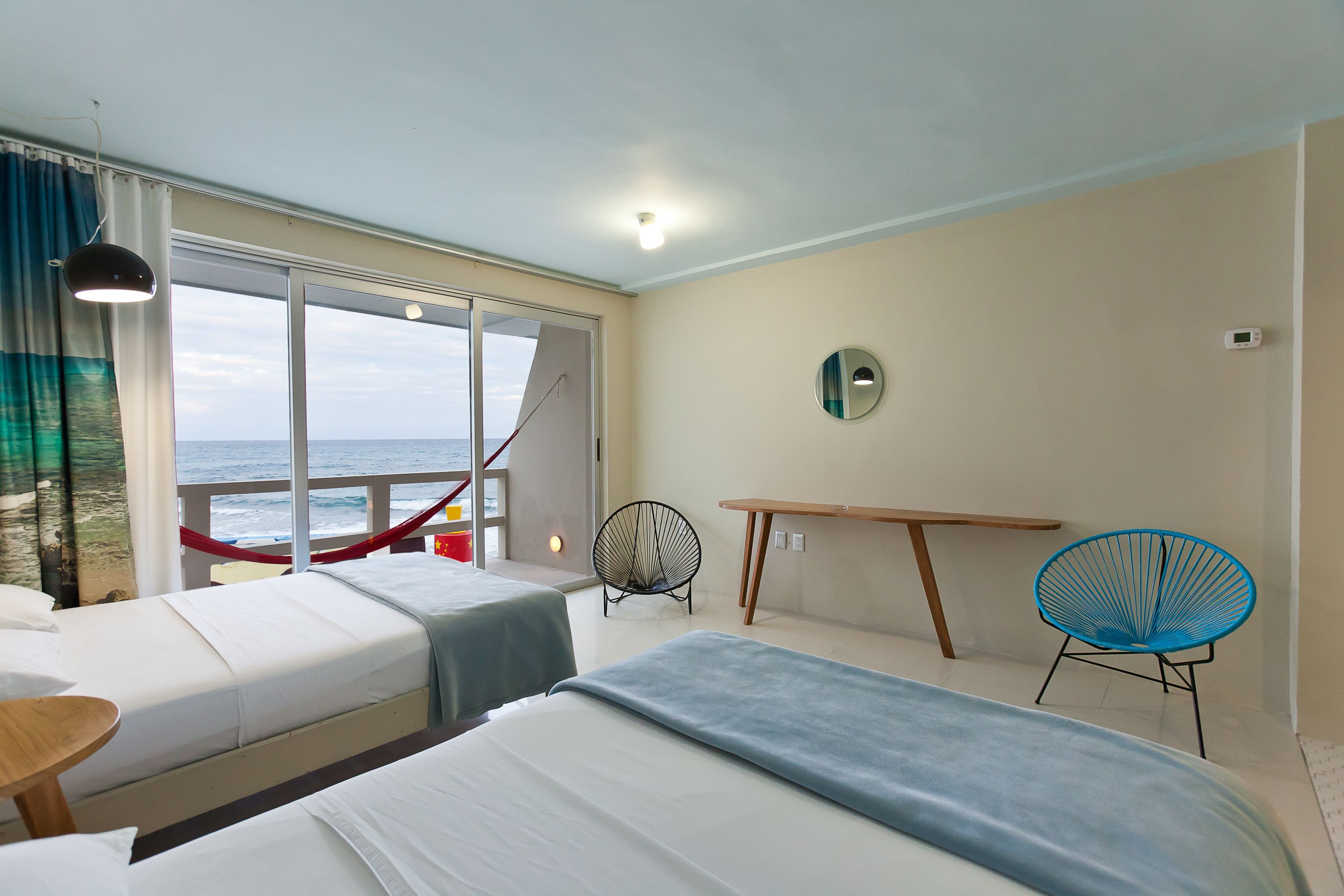 Une chambre d’hôtel avec deux lits, une table, une chaise, une fenêtre donnant sur la mer et un luminaire au plafond.