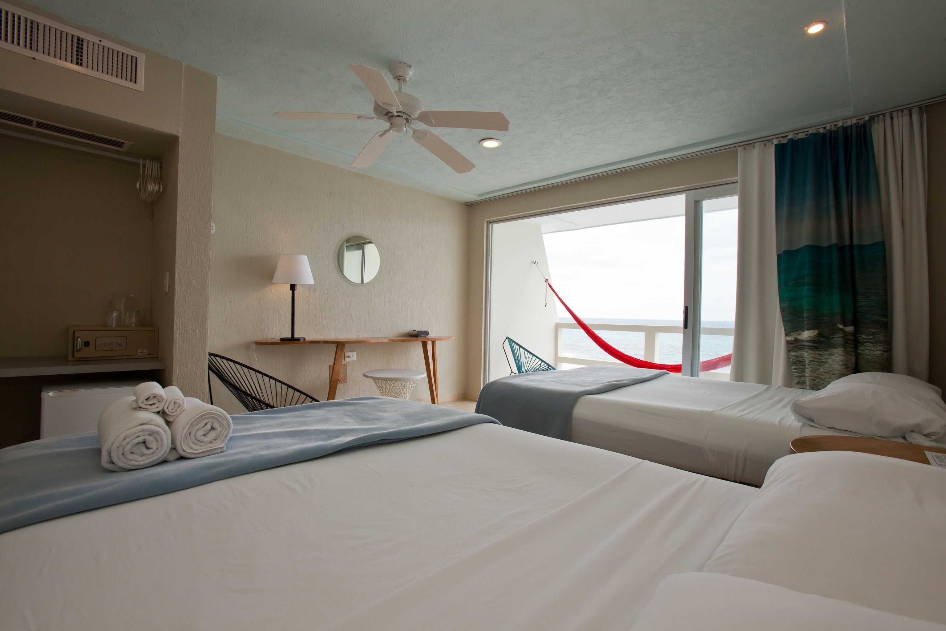 Hotelkamer met twee bedden, een plafondventilator, een bureau met stoel, een raam met gordijnen en uitzicht op zee.
