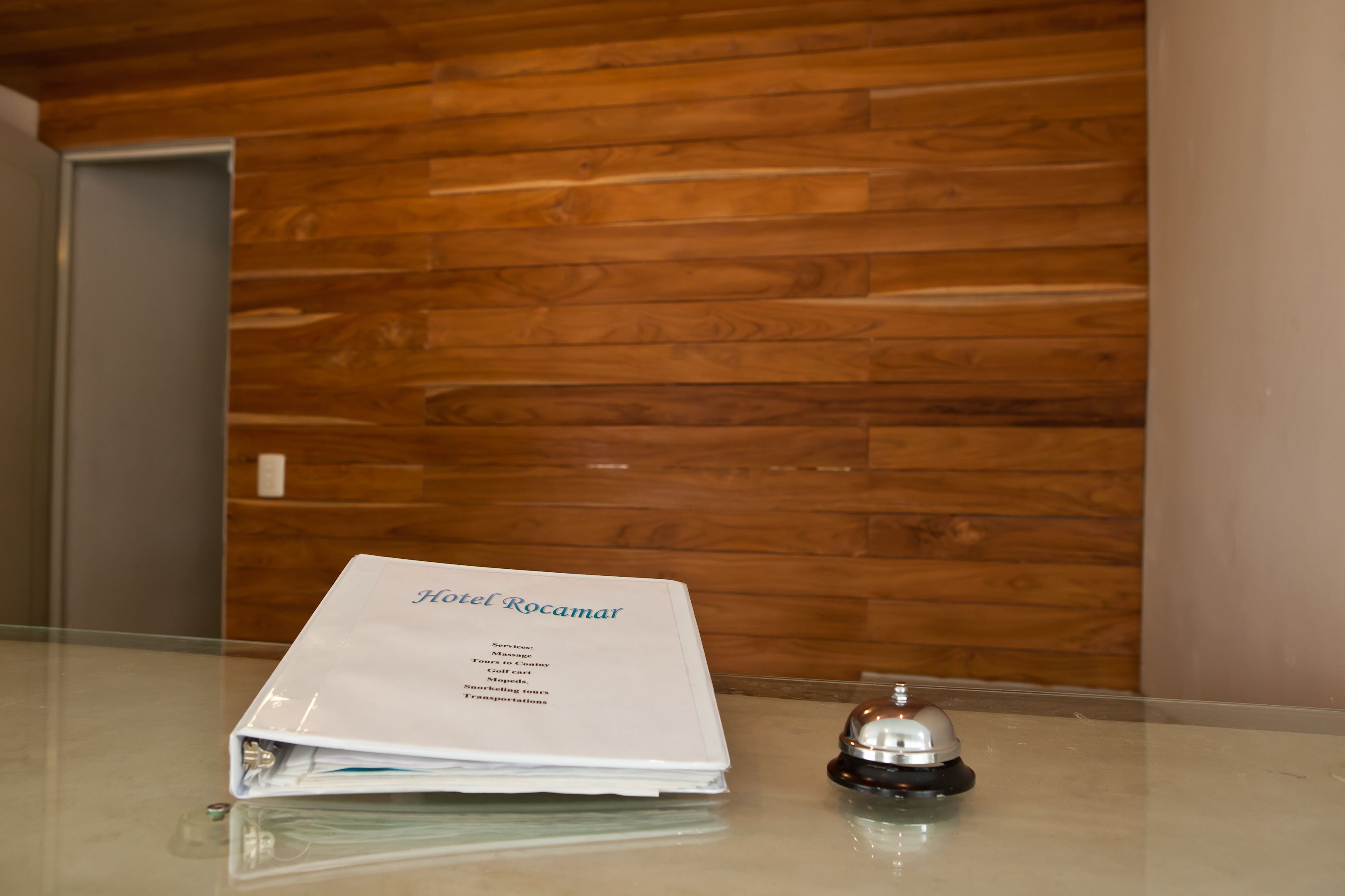 A hotel brochure titled "Hotel Rocamar" on a glass table with a wooden wall background.