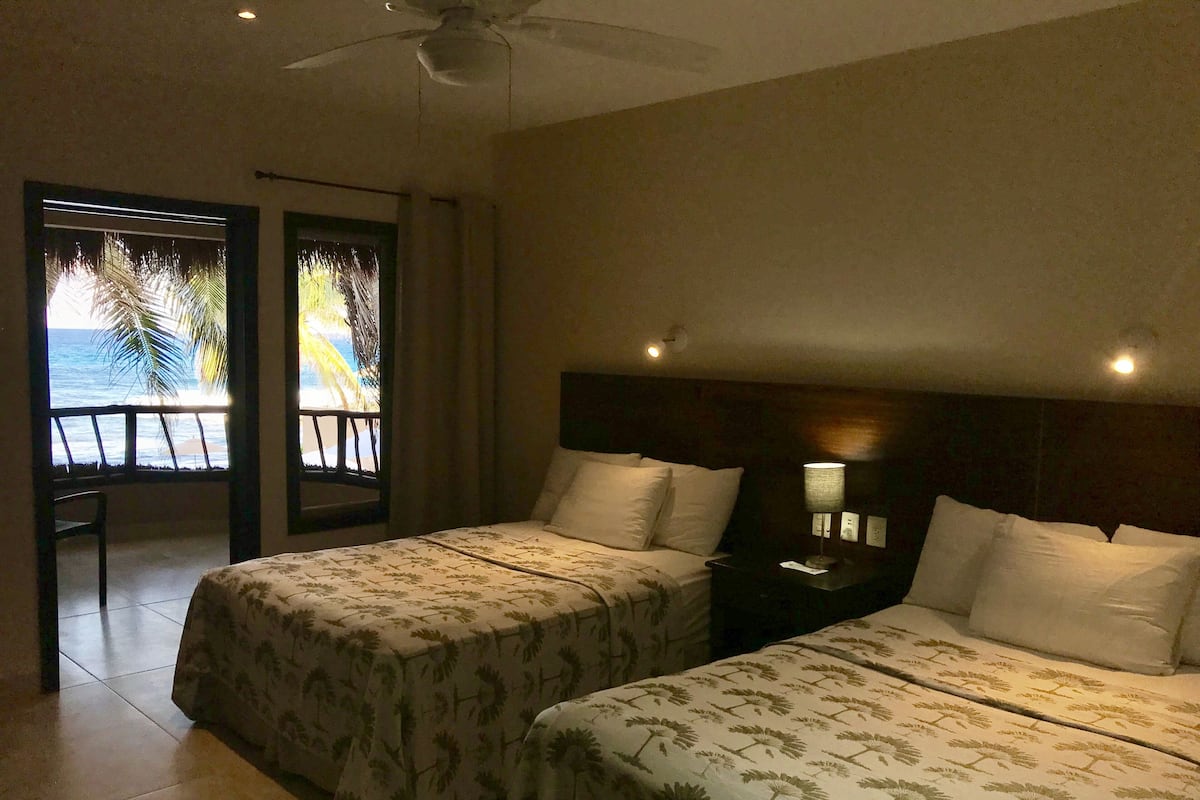 A hotel room with two beds, a balcony with a view of the ocean, and a ceiling fan.