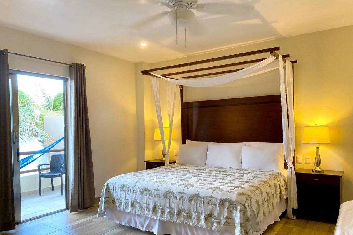 A spacious bedroom with a large bed, a canopy, a nightstand with a lamp, and a view of a balcony with a chair.