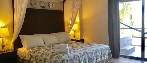 Un dormitorio con cama con dosel, dos mesitas de noche con lámparas, un ventanal con cortinas y vistas al exterior.