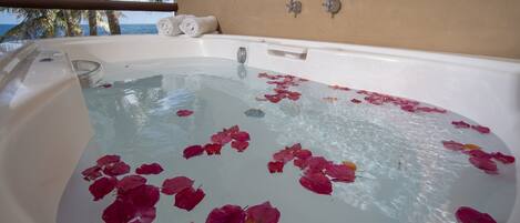 A hot tub with rose petals floating on the water, a towel, and a view of the ocean and palm trees.