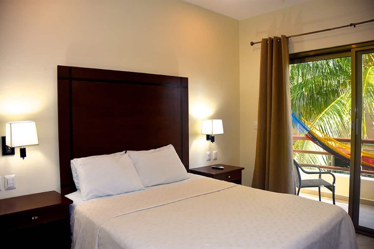 A hotel room with a large bed, two bedside tables with lamps, a chair by the window, and a view of palm trees.