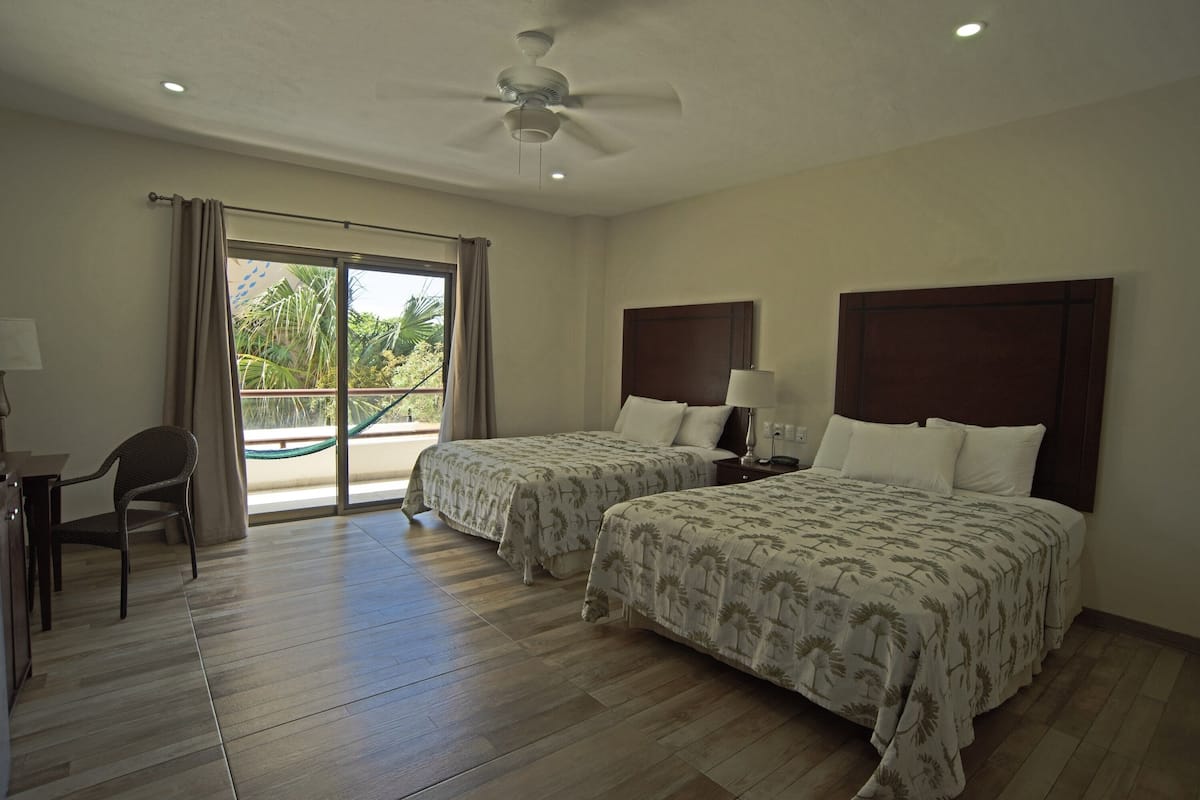 A hotel room with two beds, a desk, a chair, a ceiling fan, and a view of palm trees through the window.