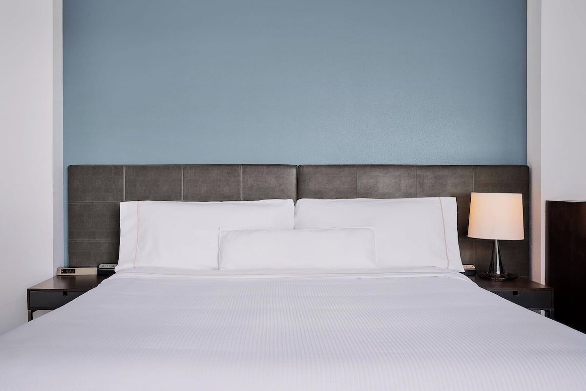 A neatly made bed with white linens and a dark headboard, a bedside lamp, and a small table with a clock.