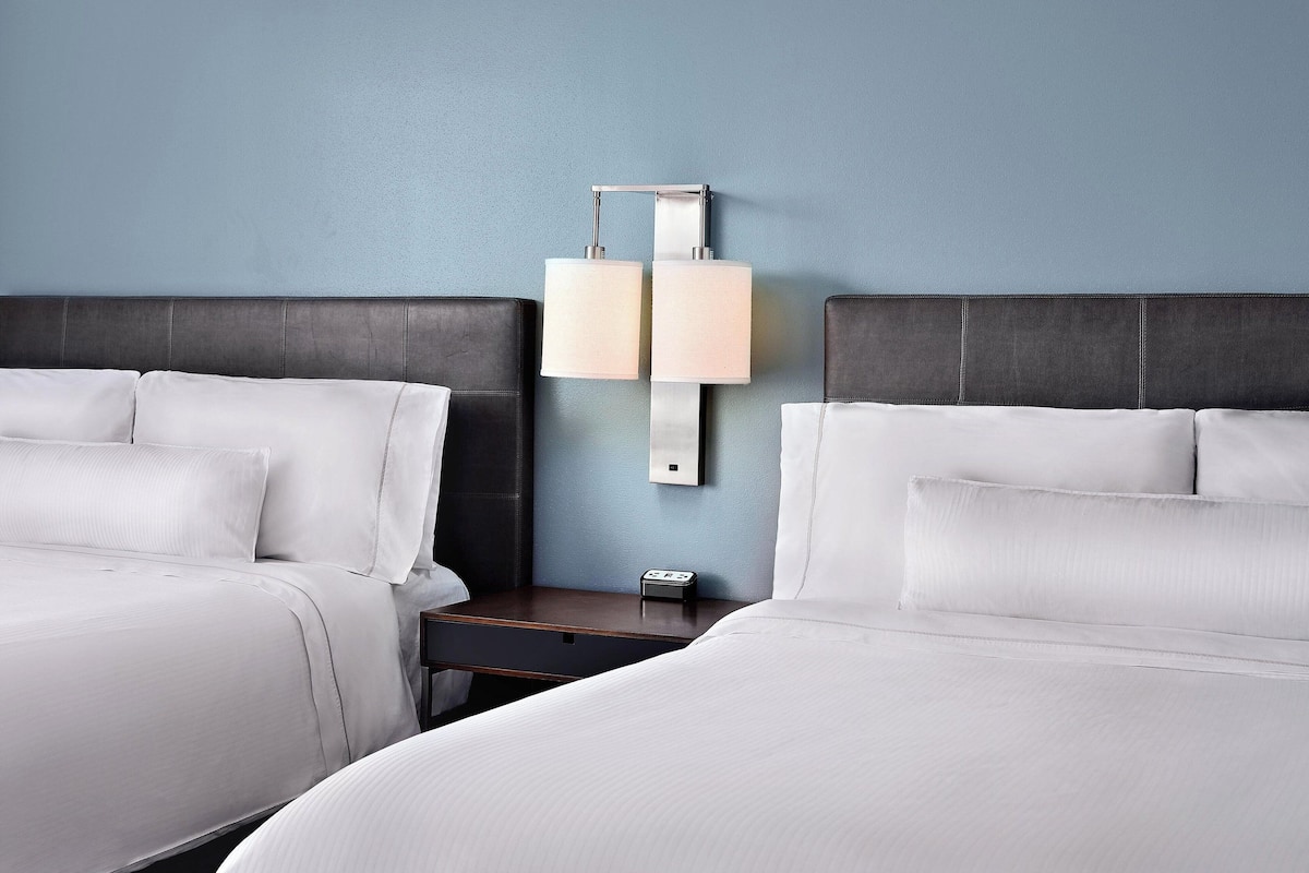 A hotel room with two beds, a nightstand, a wall-mounted lamp, and a small table.