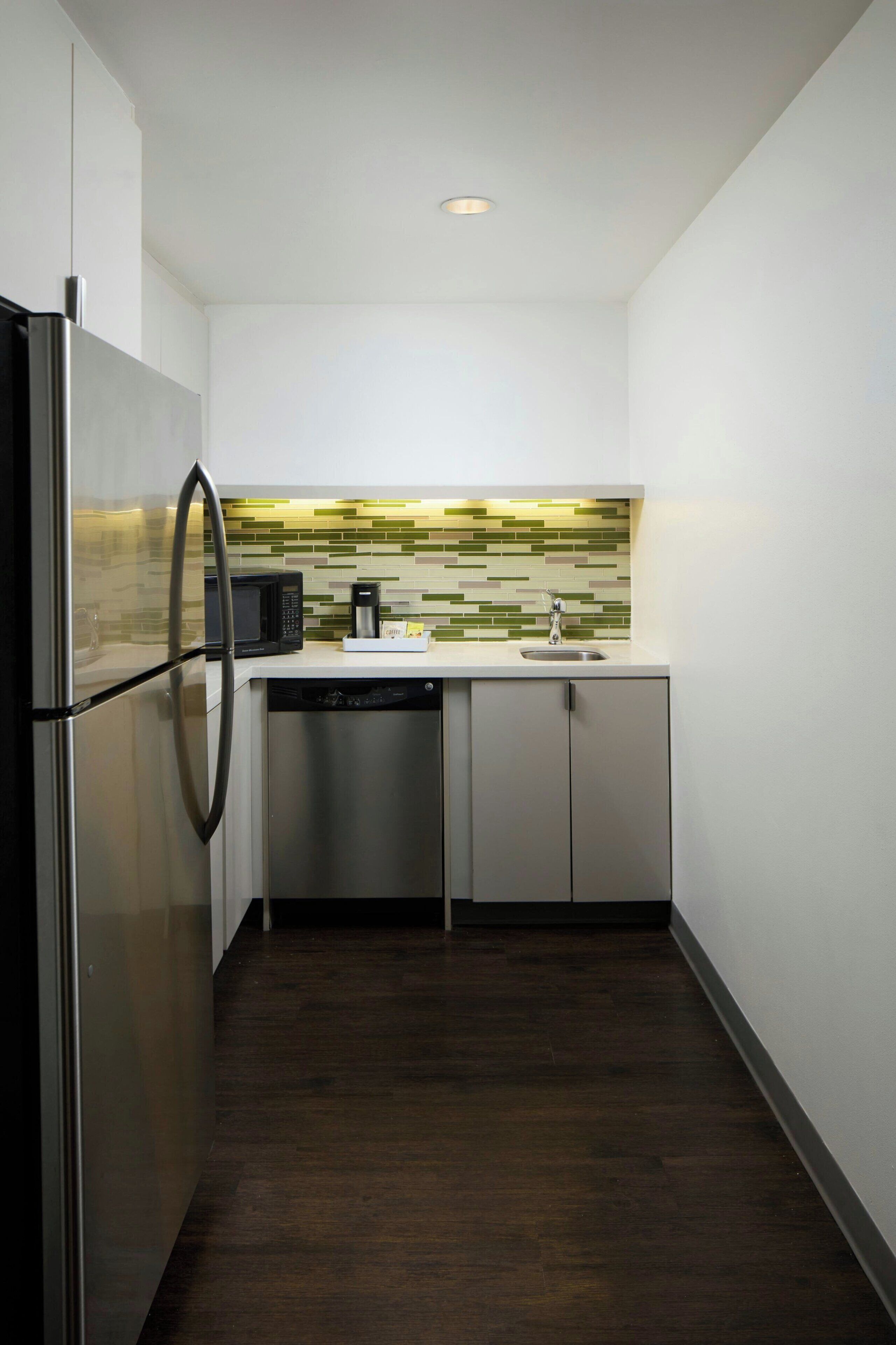 A compact kitchen with a stainless steel refrigerator, a microwave, a dishwasher, and a tiled backsplash.