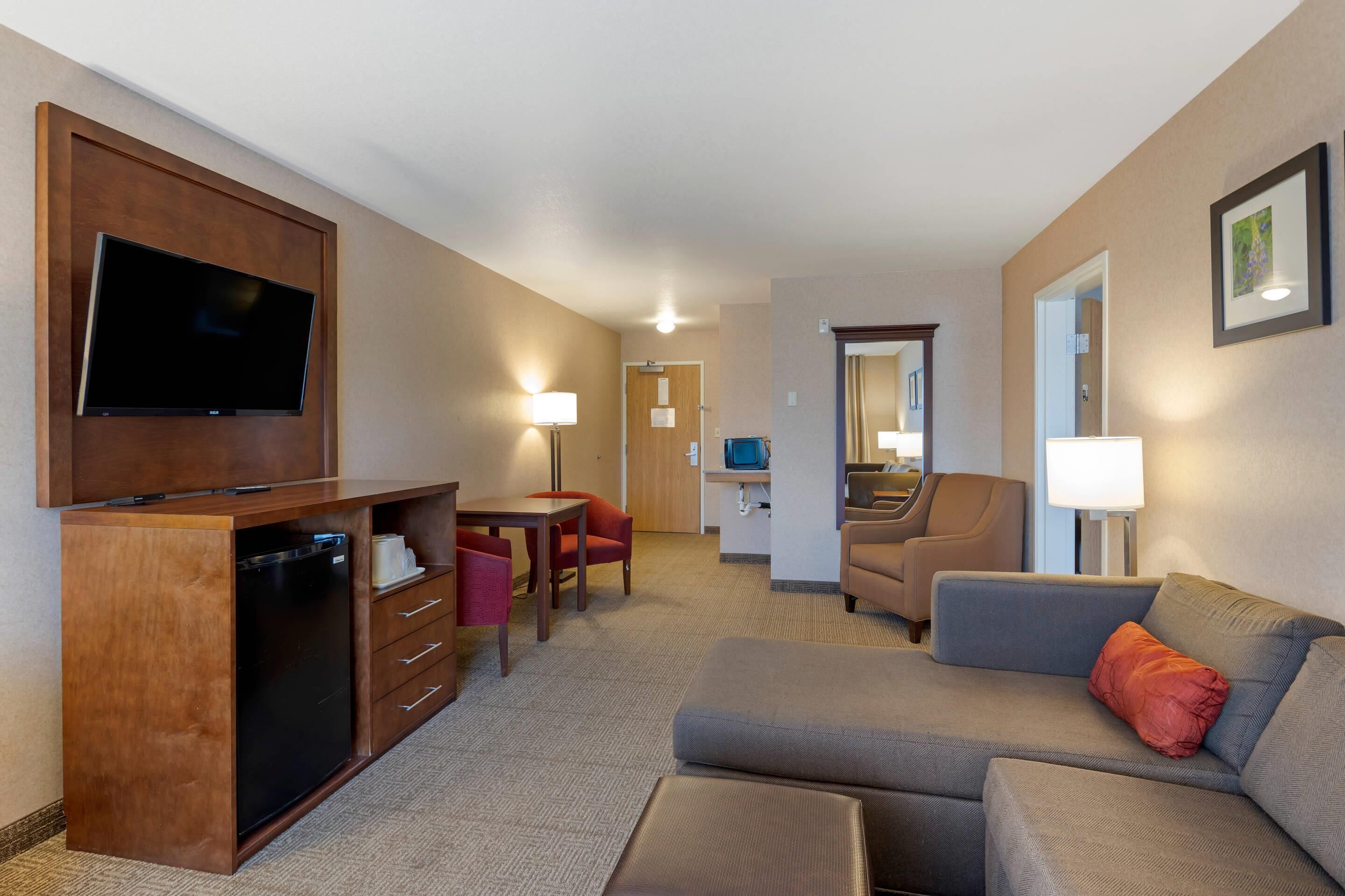 A hotel room with a flat-screen TV mounted on a wooden cabinet, a sofa, a dining table with chairs, and a kitchenette area.