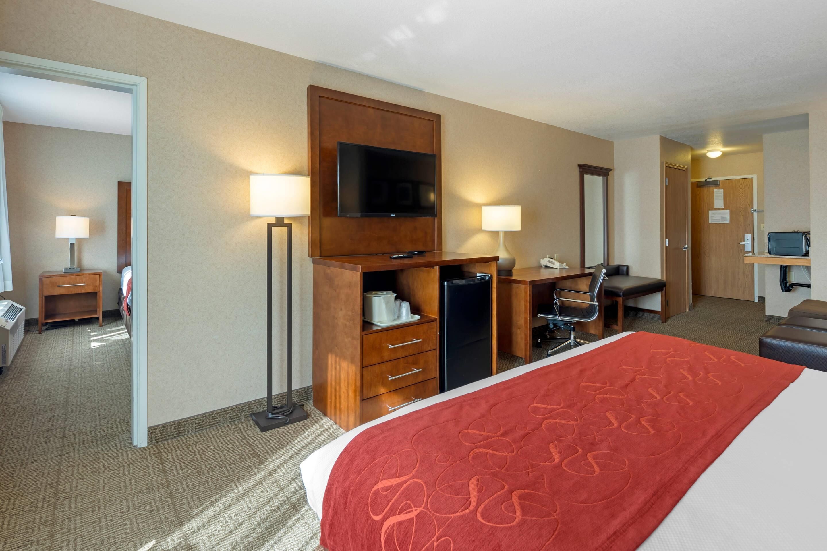 Een hotelkamer met een bed, een bureau met stoel, een televisie, een bureaulamp, een koelkast en een spiegel.