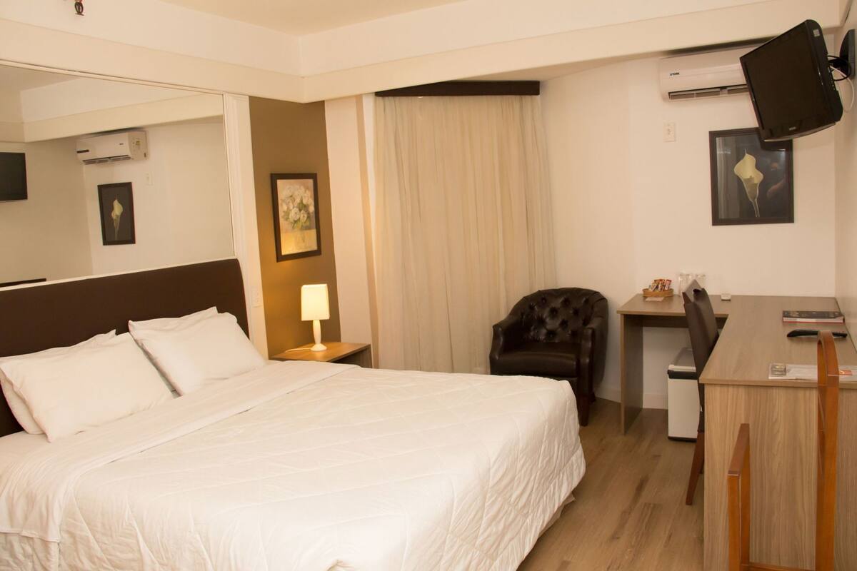 A hotel room with a bed, a desk, a chair, a lamp, and an air conditioning unit.