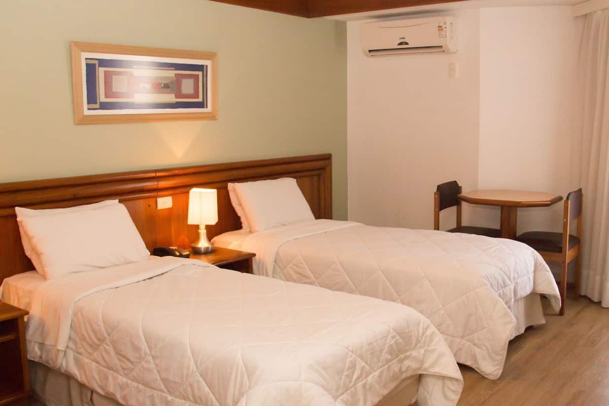 A hotel room with two beds, a nightstand with a lamp, a chair, and an air conditioning unit.