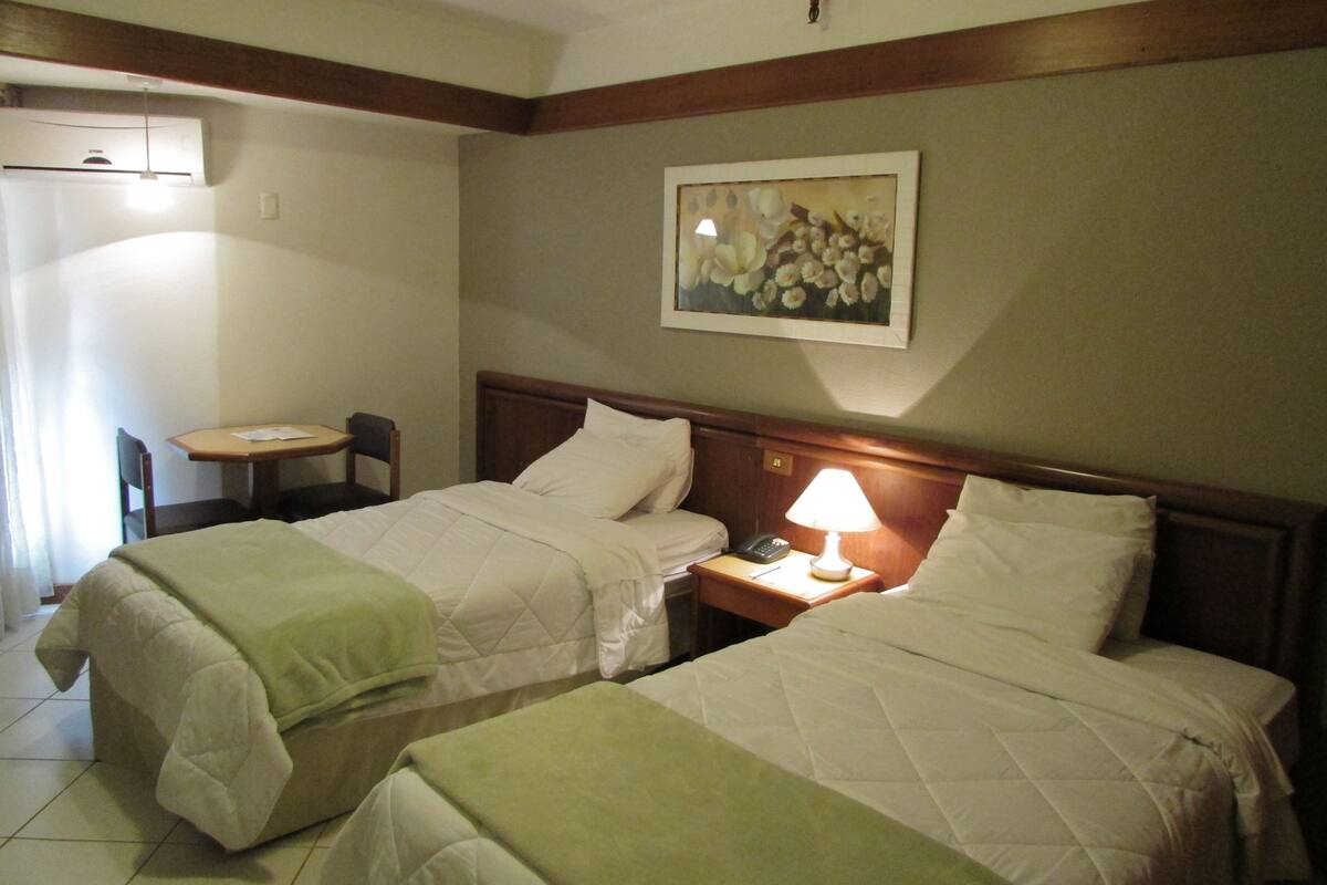 A hotel room with two beds, a small table, a chair, a lamp, and a painting on the wall.