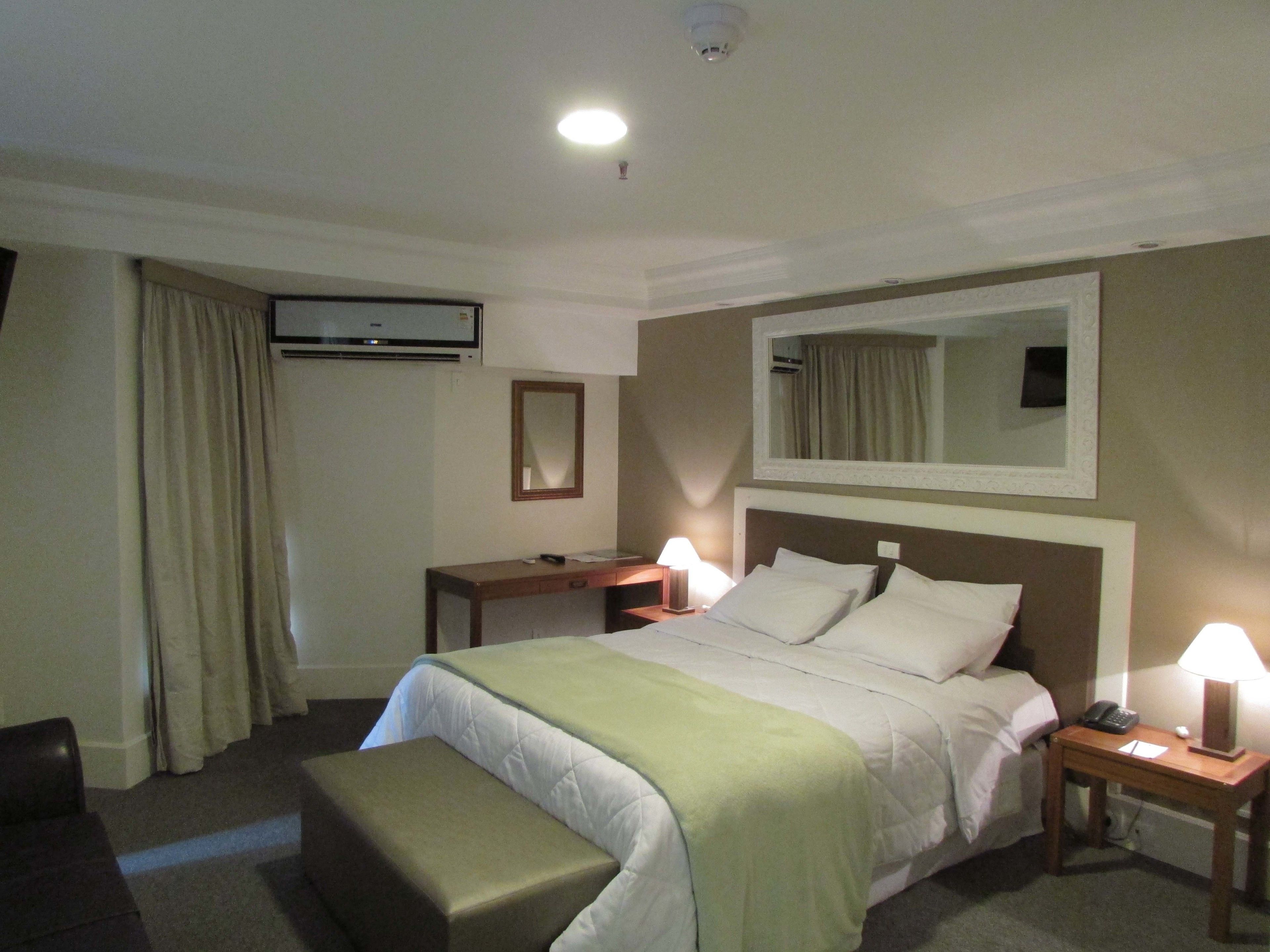 A hotel room with a bed, bedside tables, a desk, a chair, a mirror, and an air conditioning unit.