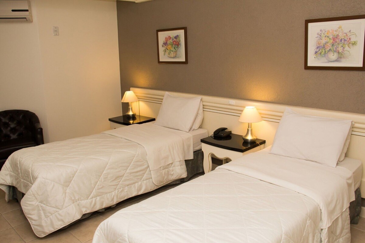 Habitación de hotel con dos camas, mesitas de noche con lámparas, una silla y dos cuadros enmarcados con motivos florales en la pared.
