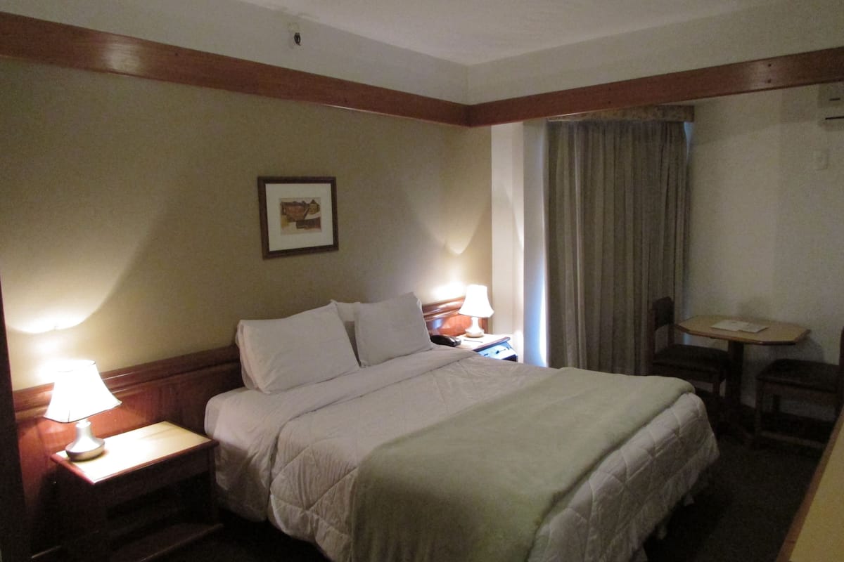 A hotel room with a bed, bedside tables, a chair, a small table, and a picture on the wall.