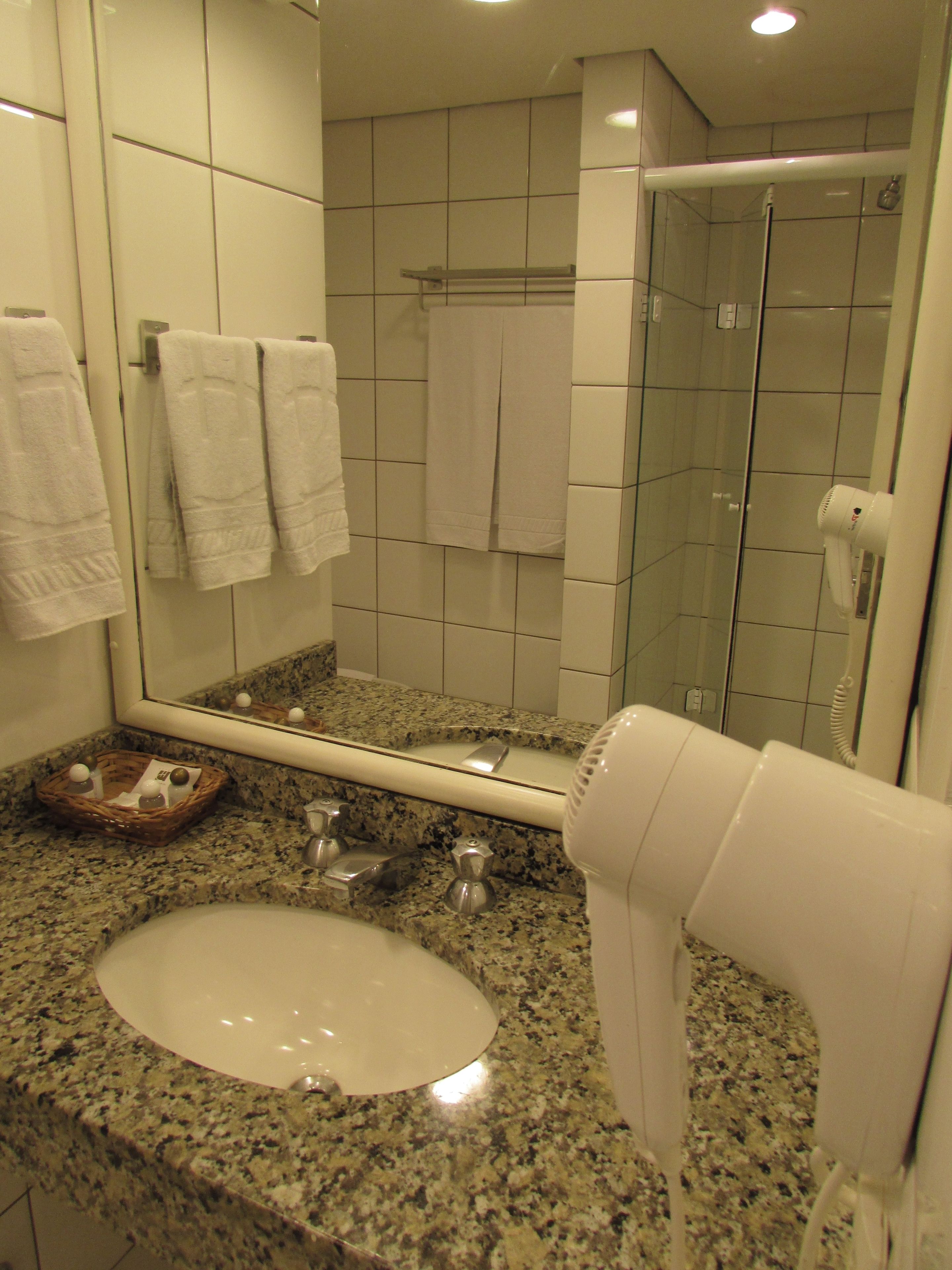 Un baño con encimera de granito, lavamanos, espejo, secador de pelo y cabina de ducha de vidrio.