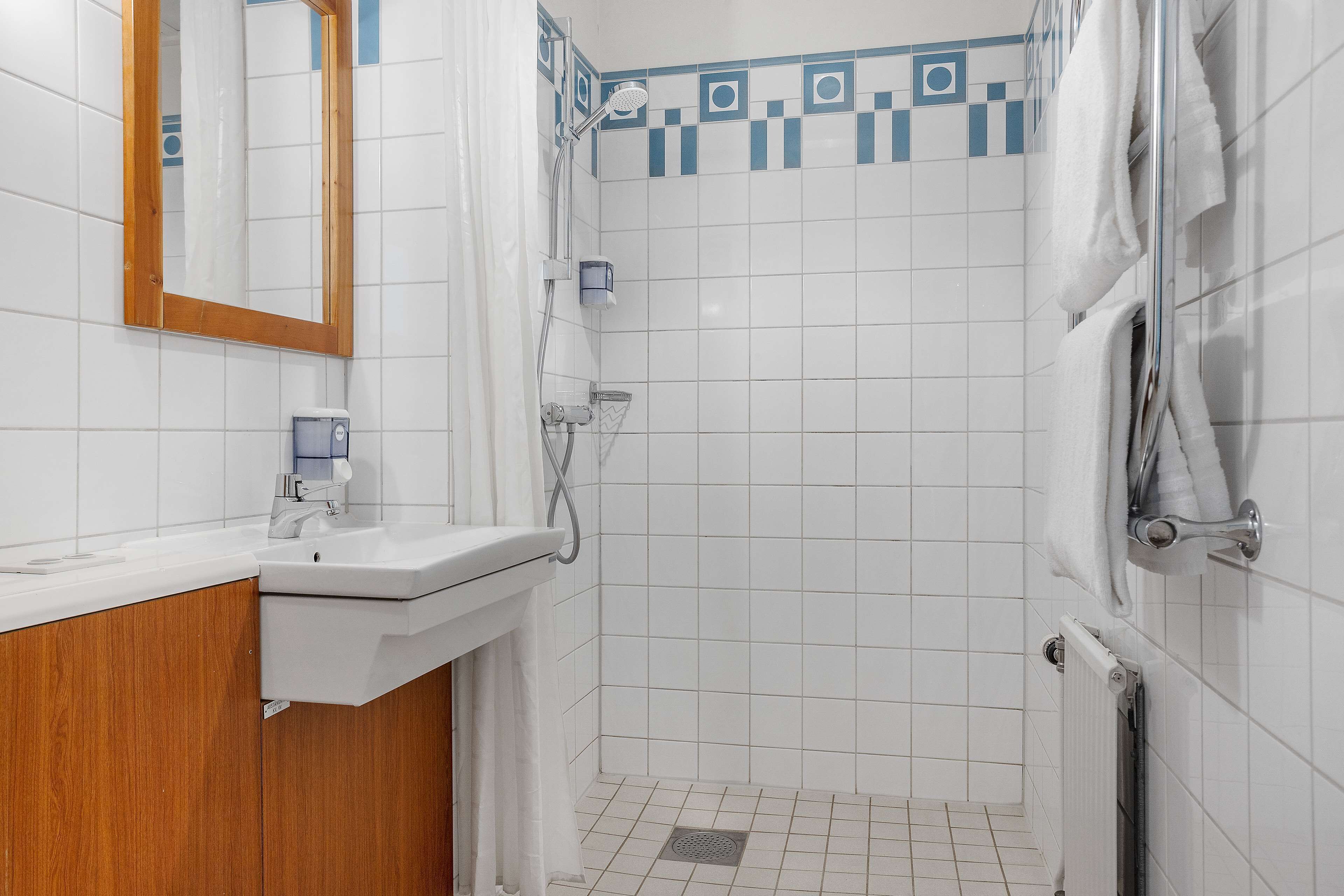 A bathroom with white tiles, a wooden vanity, a shower with a glass door, and a towel rack.