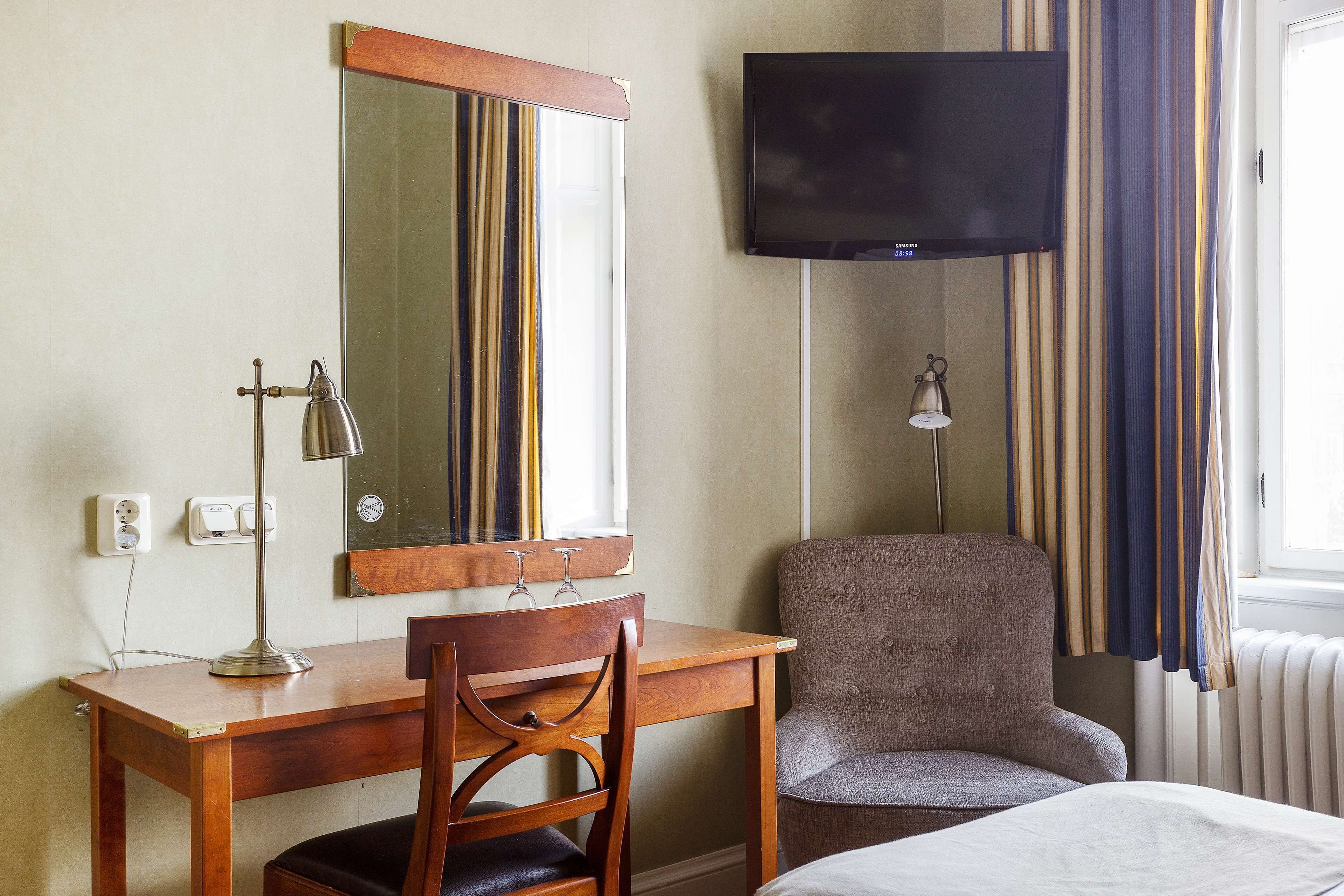 A hotel room with a wooden desk, a chair, a bed, a television, and a window with curtains.
