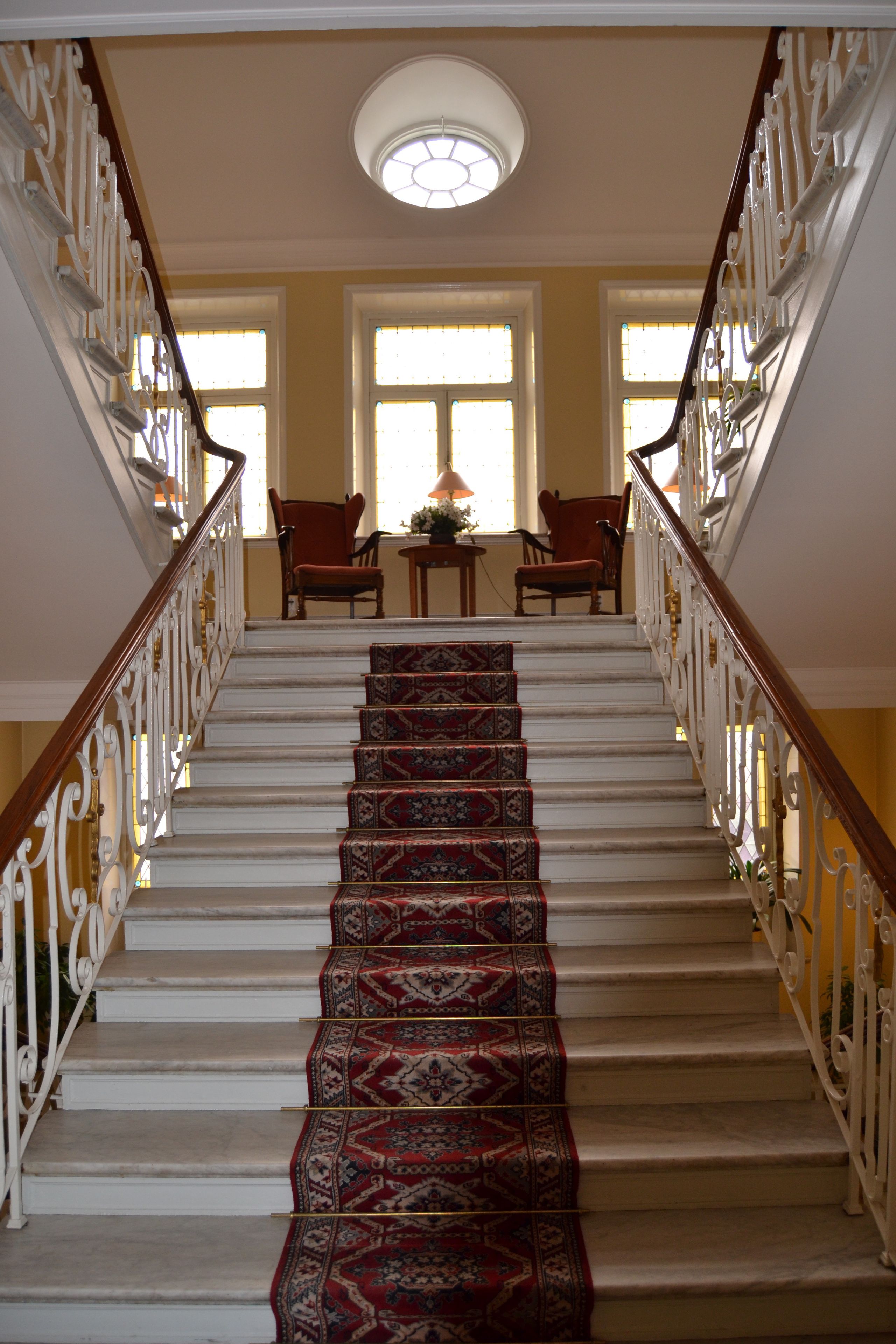 A grand staircase with ornate railings and a patterned carpet leading to a landing with two chairs and a table.