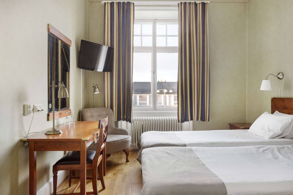A hotel room with a wooden desk, two beds, a TV, and a window with curtains.