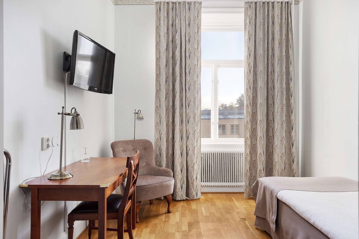 A room with a wooden dining table, a single chair, a flat-screen TV mounted on the wall, and a bed with a white bedspread.
