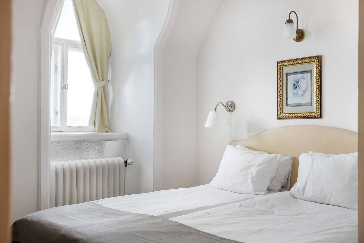 A bedroom with a bed, a window with curtains, a radiator, a wall-mounted light fixture, and a framed picture.