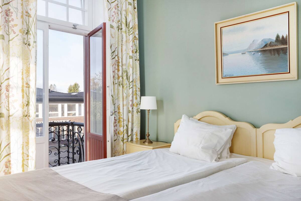 A hotel room with a bed, a nightstand with a lamp, a painting on the wall, and a view of a balcony with a railing.