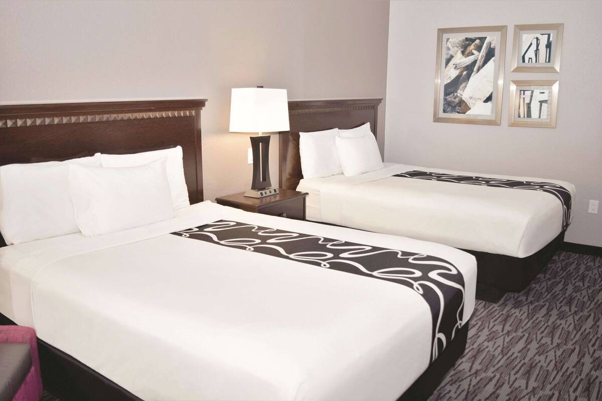 Ein Hotelzimmer mit zwei Betten, einem Nachttisch mit Lampe und gerahmten Kunstwerken an der Wand.