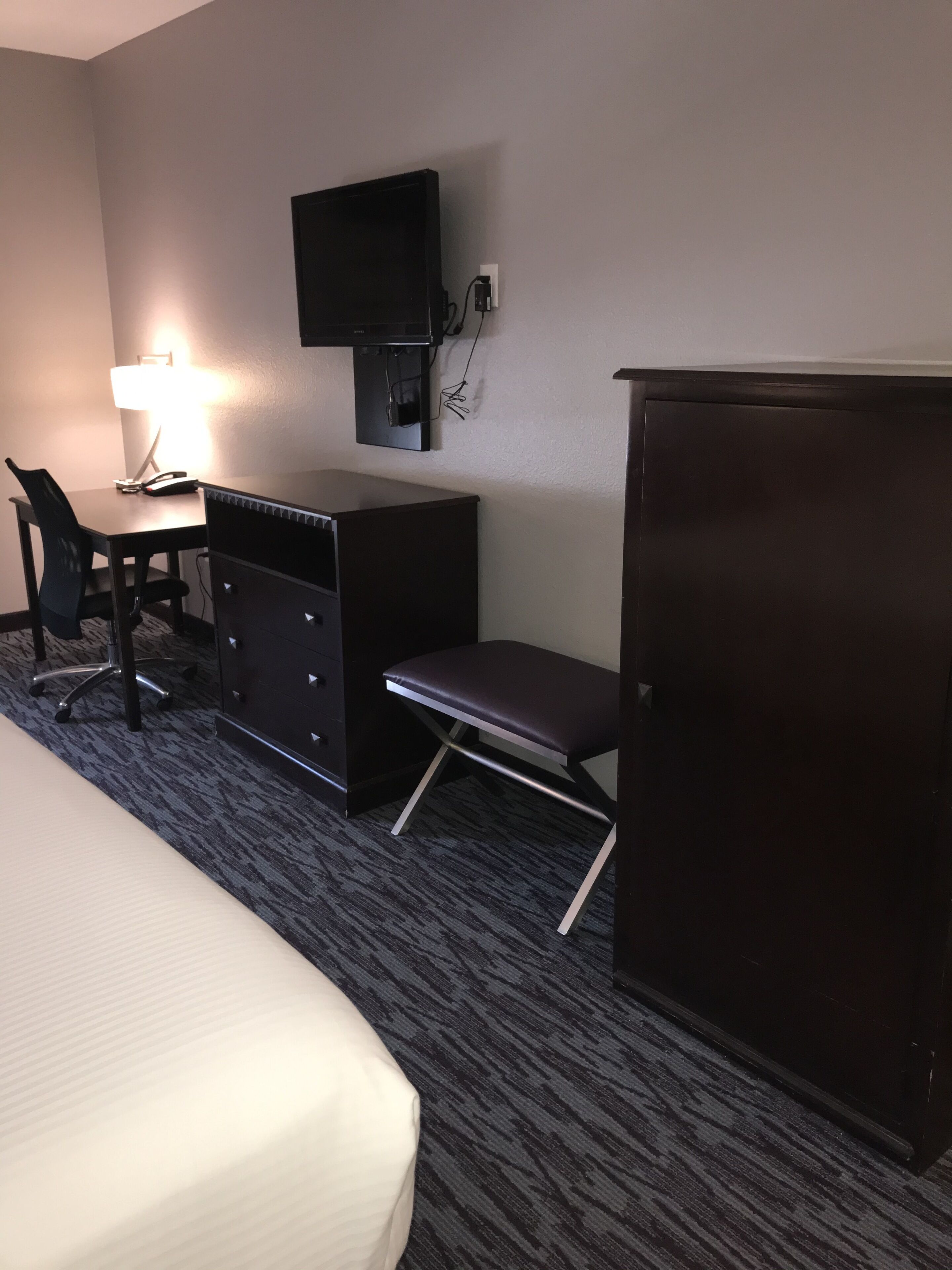 A hotel room with a bed, a desk with a lamp, a chair, a television mounted on the wall, and a wardrobe.