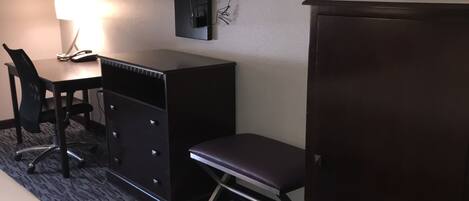 A hotel room with a bed, a desk with a lamp, a chair, a television mounted on the wall, and a wardrobe.