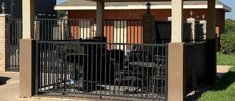 A covered outdoor seating area with a metal fence, a gazebo structure, and a building in the background.