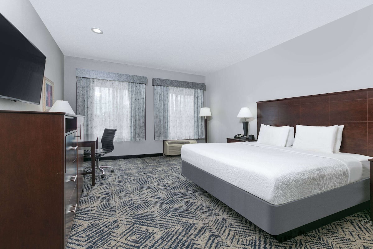 A hotel room with a large bed, a desk with a chair, a TV, and two windows with curtains.