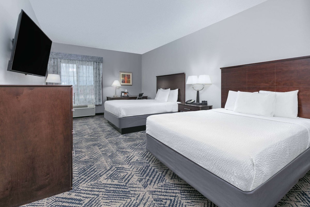 A hotel room with two beds, a flat-screen TV, a desk with a lamp, and a window with curtains.