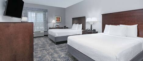 A hotel room with two beds, a flat-screen TV, a desk with a lamp, and a window with curtains.