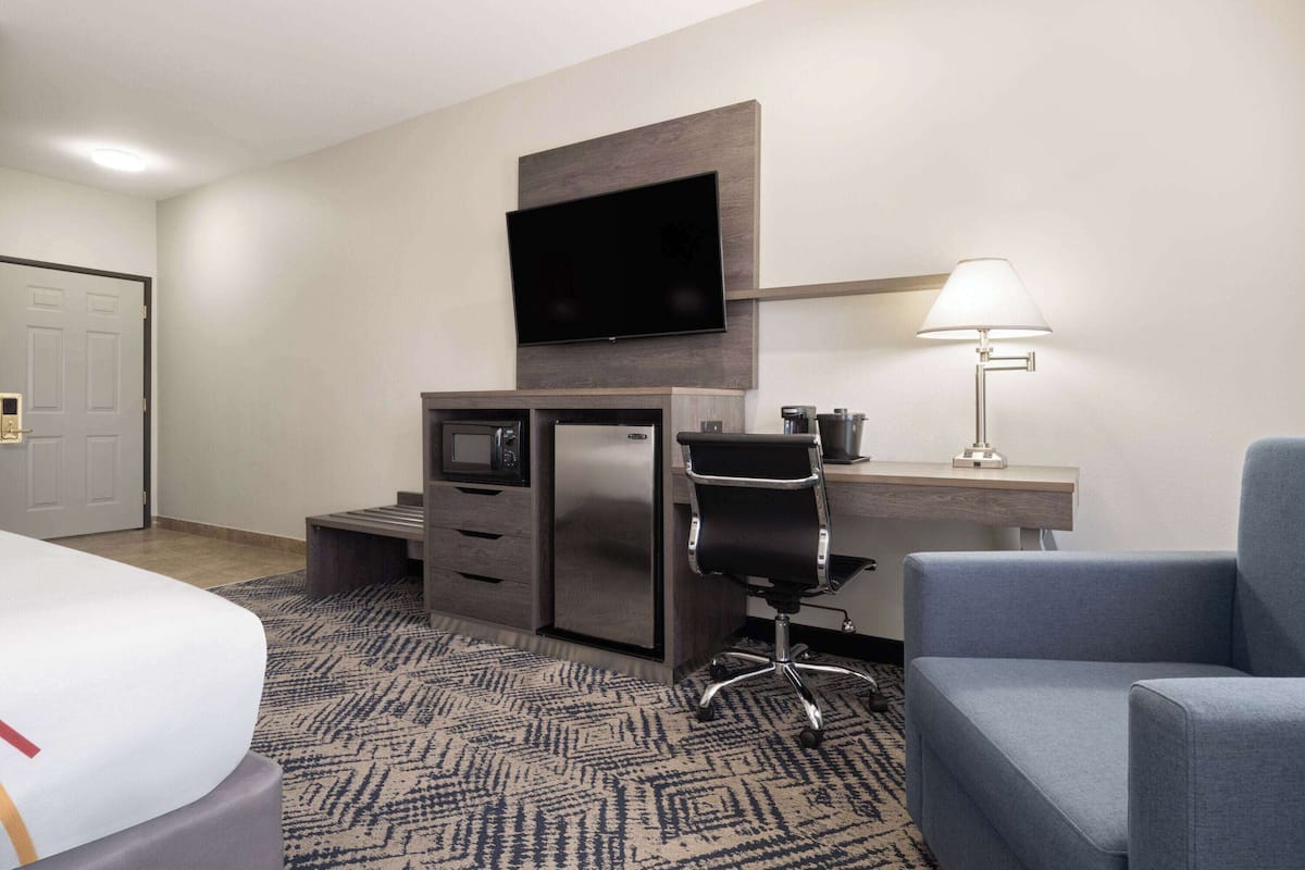 A hotel room with a bed, a desk with a chair, a television, a lamp, and a minibar.